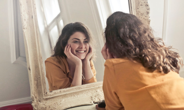 Curly-haired brunette young woman wearing a yellow shirt looks into a mirror smiling as she checks for gum disease